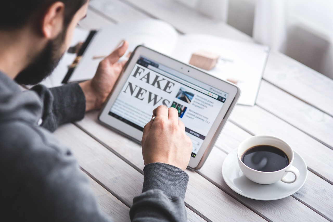Uomo che legge notizie su un tablet con la scritta ‘Fake News’ e una tazza di caffè accanto.